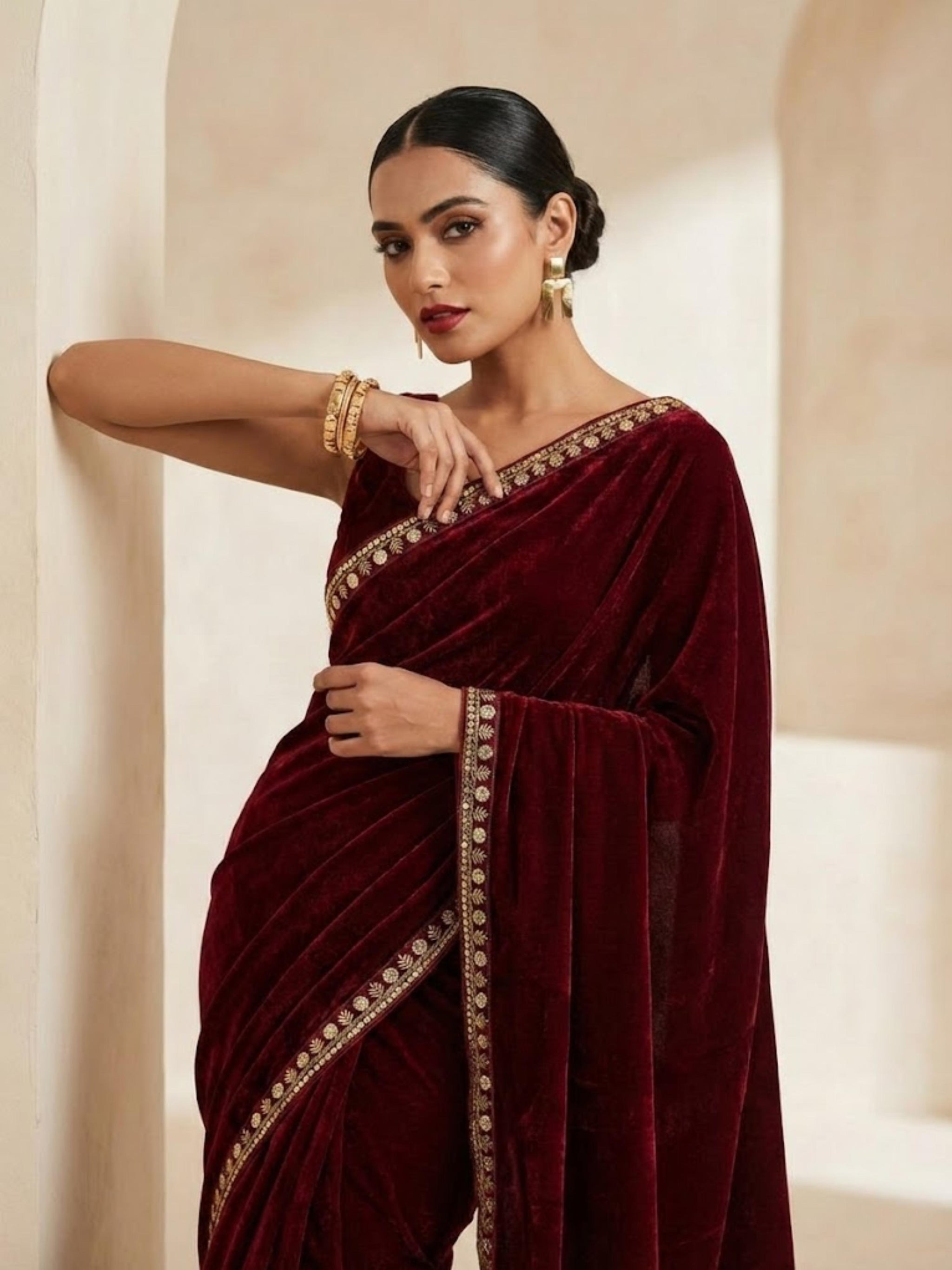Woman wearing a maroon saree with gold embroidery against a beige background