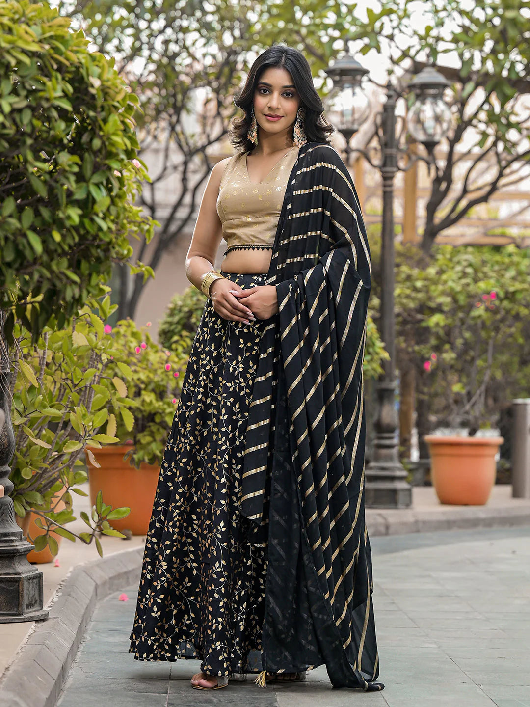 Woman in a black and gold saree standing outdoors with greenery in the background