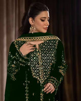 Woman wearing a dark green embroidered traditional outfit with gold details against a brown curtain background.